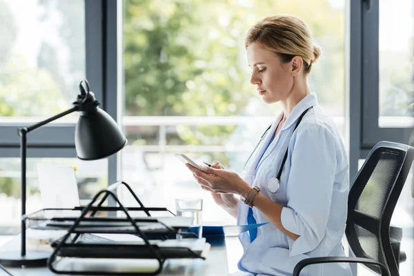 Боковой вид женщины-врача в белом пальто, использующей смартфон за столом в офисе — стоковое фото