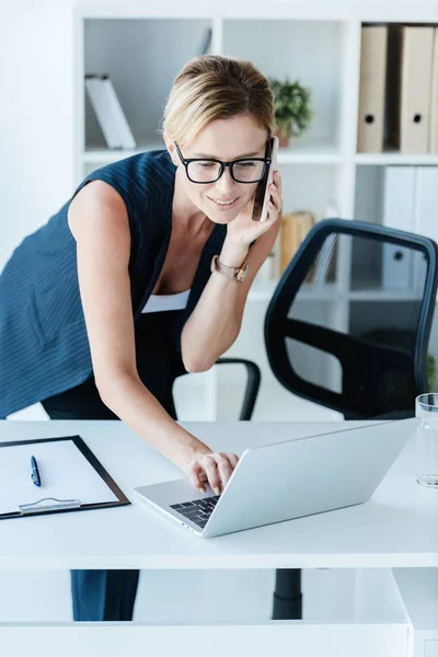 Приваблива бізнес-леді в окулярах розмовляє на смартфоні і використовує ноутбук в офісі — стокове фото