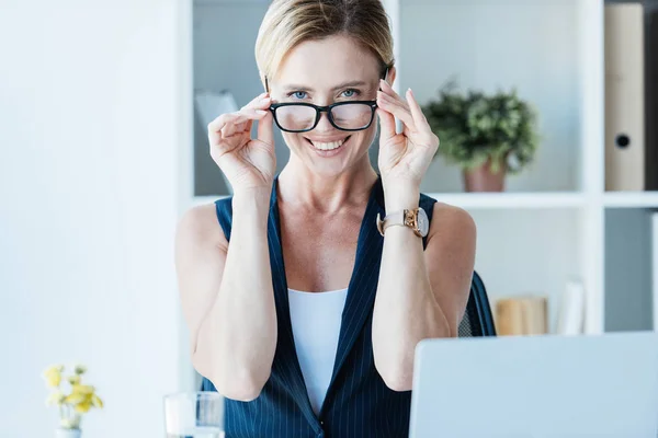Усміхнена доросла бізнес-леді регулює окуляри і дивиться на камеру в офісі — стокове фото