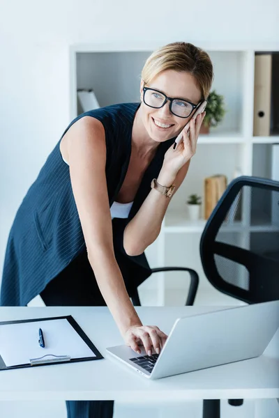 Donna d'affari adulta in occhiali che parla su smartphone e utilizza il computer portatile in ufficio — Foto stock
