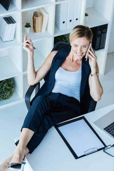 Высокий угол зрения улыбающаяся деловая женщина с ногами на столе говорить на смартфоне в офисе — стоковое фото
