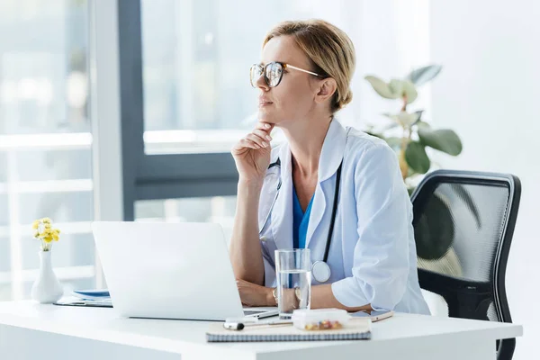 Médico femenino reflexivo en gafas que se sienta en la mesa con el ordenador portátil en la oficina - foto de stock