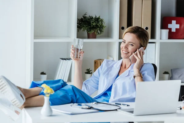 Medico donna sorridente con le gambe sulla tavola acqua potabile e parlando sullo smartphone in ufficio — Foto stock