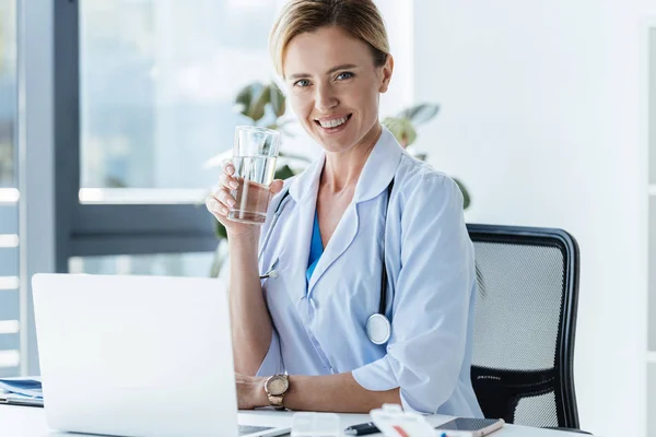 Feliz médico femenino sosteniendo un vaso de agua y sentado en la mesa con el ordenador portátil en la oficina - foto de stock