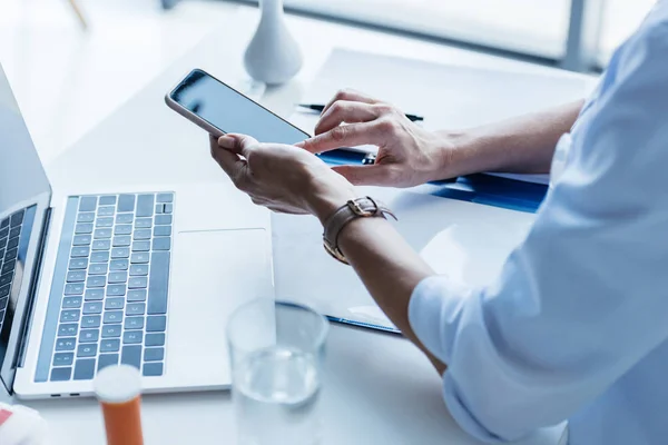 Vista parcial del médico femenino usando el teléfono inteligente en la mesa con el ordenador portátil en la oficina - foto de stock