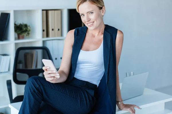 Femme d'affaires adulte heureuse assise sur la table tout en utilisant un smartphone au bureau — Photo de stock