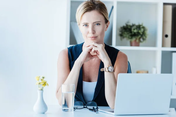 Retrato de mujer de negocios adulta mirando a la cámara y sentado en la mesa con el ordenador portátil en la oficina - foto de stock