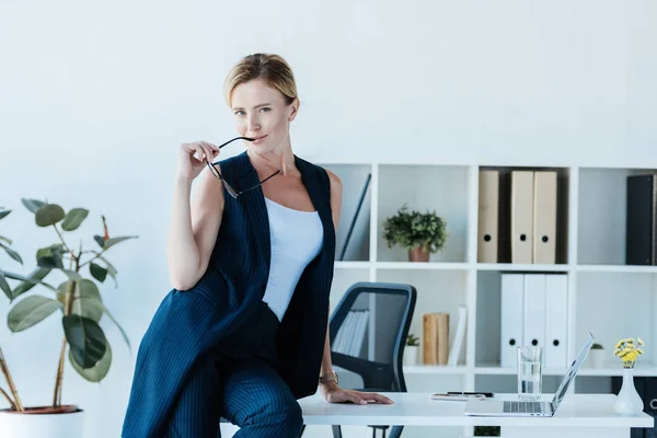 Красива доросла бізнес-леді тримає окуляри і позує на столі з ноутбуком в офісі — стокове фото