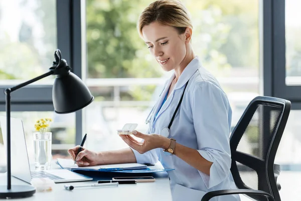 Erwachsene Ärztin schreibt in Klemmbrett und hält Tabletten am Tisch im Büro — Stockfoto