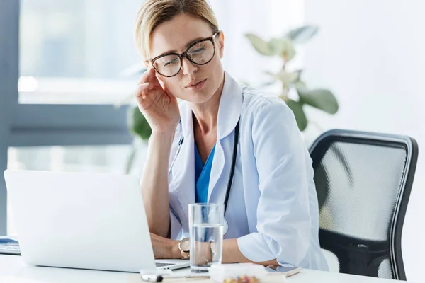 Exhausted female doctor in eyeglasses having headache at table in office — Stock Photo