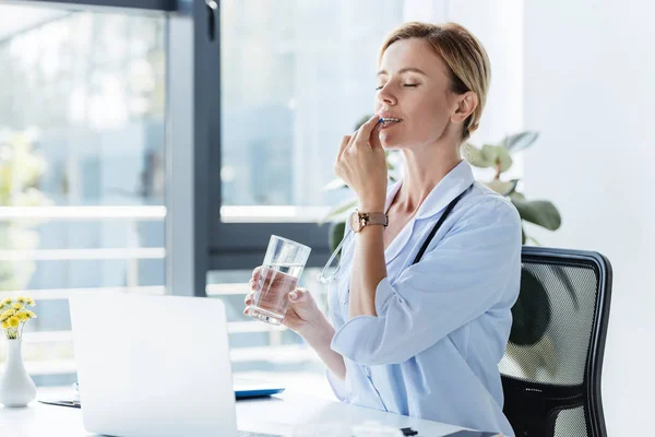 Doctora adulta en bata blanca sosteniendo vaso de agua y tomando píldora en la mesa con computadora portátil en la oficina - foto de stock