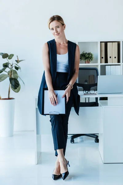 Adulto mujer de negocios de pie con portapapeles cerca de la mesa con el ordenador portátil en la oficina - foto de stock