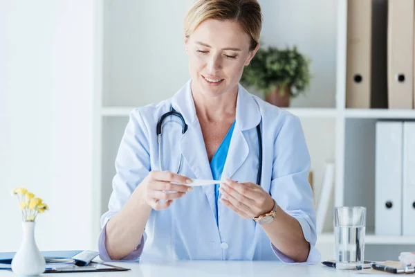 Adult female doctor in white coat checking thermometer at table in office — Stock Photo