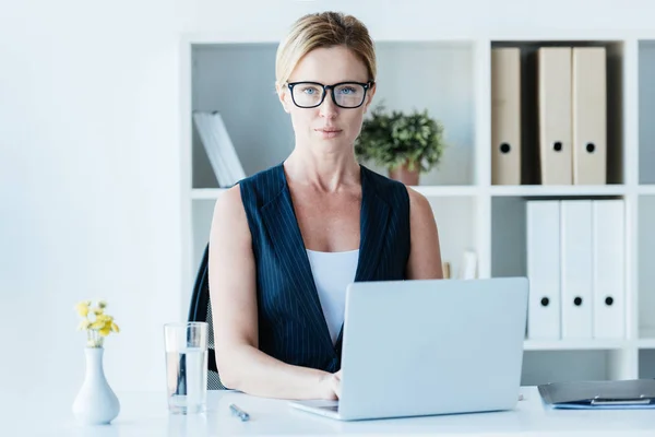 Serious adult businesswoman in eyeglasses working with laptop at table in office — Stock Photo