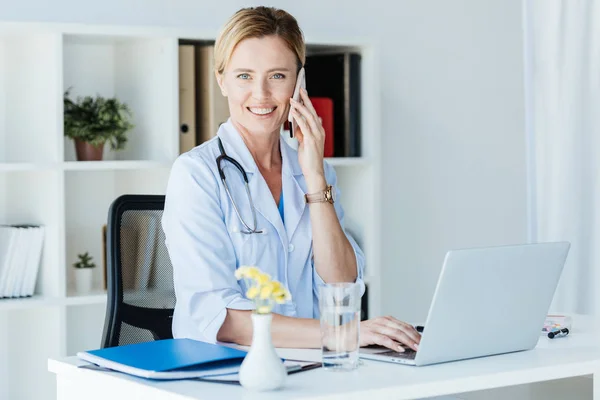 Щаслива жінка-лікар дивиться на камеру і говорить на смартфон за столом з ноутбуком в офісі — стокове фото