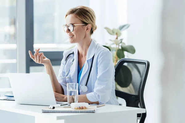 Adulto feliz mujer médico en gafas que habla en la mesa con el ordenador portátil en la oficina - foto de stock