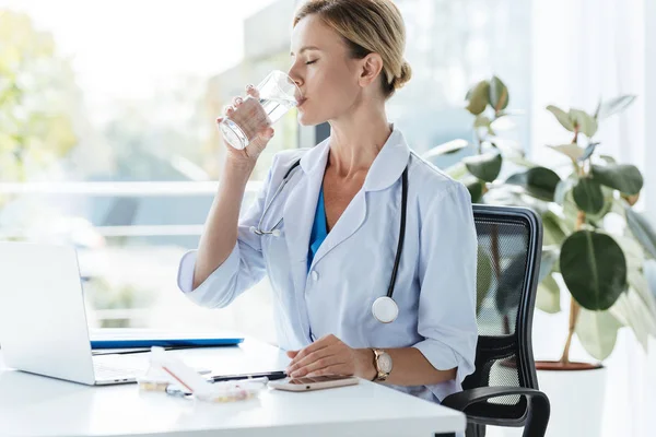 Erwachsene Ärztin in weißem Mantel mit Stethoskop über dem Hals trinkt Wasser am Tisch im Büro — Stockfoto