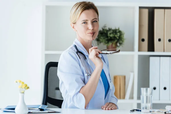 Attractive female doctor in white coat with stethoscope over neck holding eyeglasses at table in office — Stock Photo