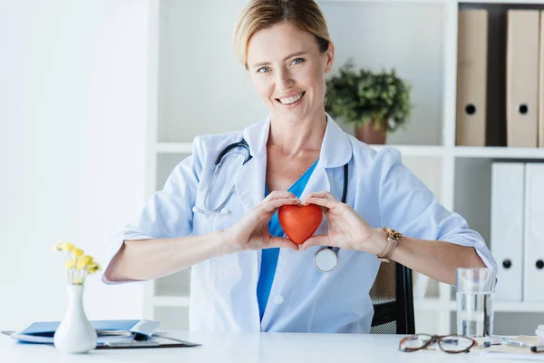 Happy female doctor in white coat showing heart symbol at table in office — Stock Photo
