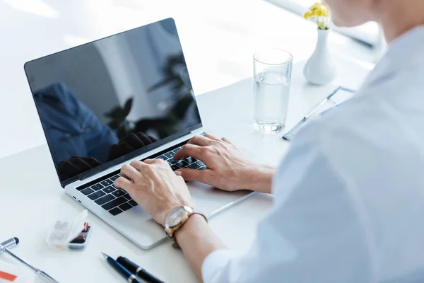 Partial view of female doctor typing on laptop with blank screen at table in office — Stock Photo