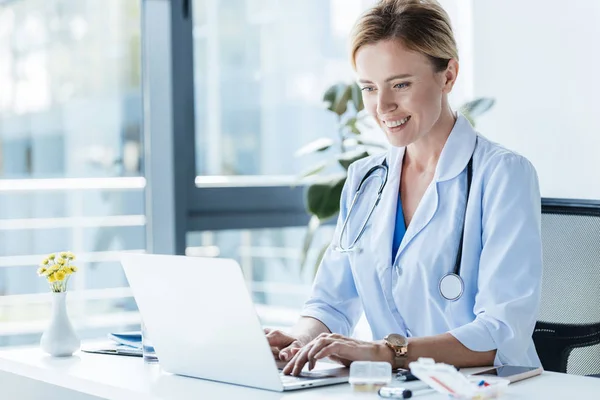 Smiling adult female doctor in white coat using laptop at table in office — Stock Photo