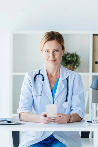 Doctora en bata blanca usando teléfono inteligente y mirando a la cámara en la mesa en la oficina - foto de stock