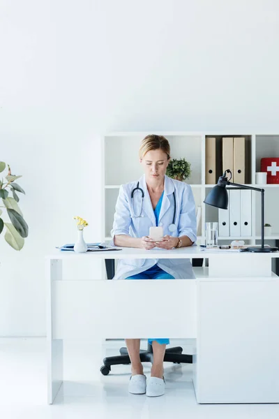 Doctora adulta en bata blanca usando teléfono inteligente en la mesa en la oficina - foto de stock