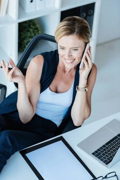 Високий кут зору усміхненої бізнес-леді розмовляє на смартфоні за столом з ноутбуком і кишені в офісі — стокове фото