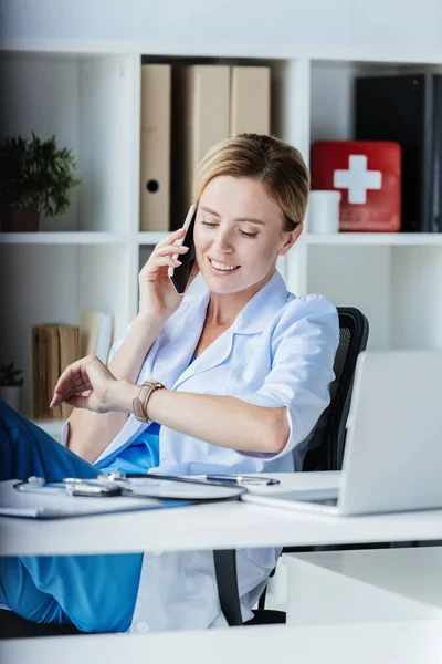 Souriant femme médecin vérifier montre-bracelet et parler sur smartphone à table avec ordinateur portable dans le bureau — Photo de stock