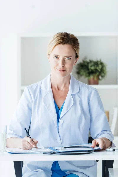 Adult female doctor in white coat writing in clipboard at table in office — Stock Photo