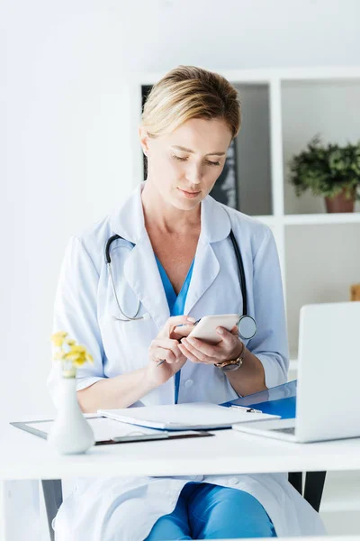 Adult female doctor using smartphone at table with laptop in office — Stock Photo