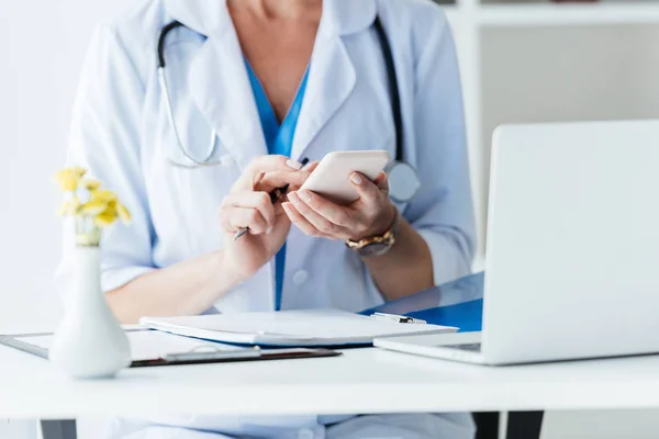 Abgeschnittenes Bild einer Ärztin mit Smartphone am Tisch mit Laptop im Büro — Stockfoto
