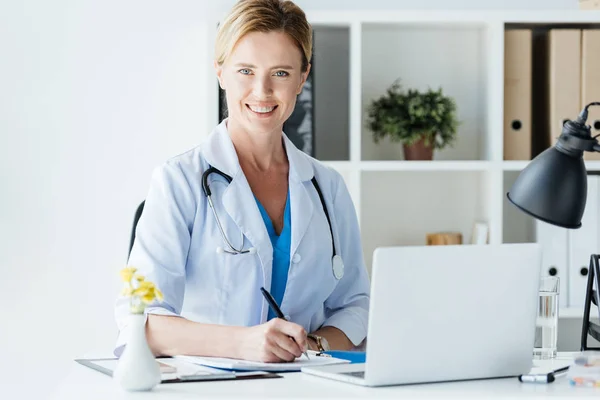 Доросла жінка-лікар зі стетоскопом над шиєю, що пише в буфері за столом з ноутбуком в офісі — стокове фото