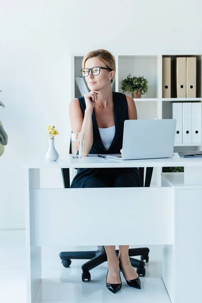 Задумчивая взрослая деловая женщина в очках сидит за столом с ноутбуком в офисе — стоковое фото