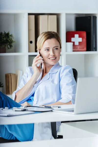 Female doctor looking away and talking on smartphone at table with laptop in office — Stock Photo