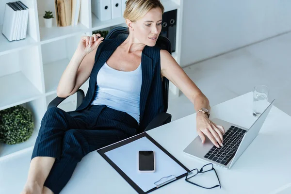 Высокий угол обзора деловой женщины, использующей ноутбук за столом со смартфоном и буфером обмена в офисе — стоковое фото