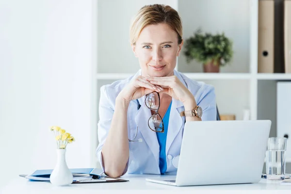 Lächelnde Ärztin mit Brille blickt auf Kamera am Tisch mit Laptop im Büro — Stockfoto