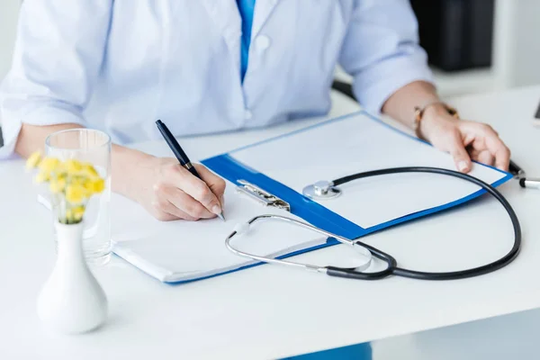 Imagem cortada de médico feminino escrevendo na área de transferência à mesa com estetoscópio no escritório — Fotografia de Stock