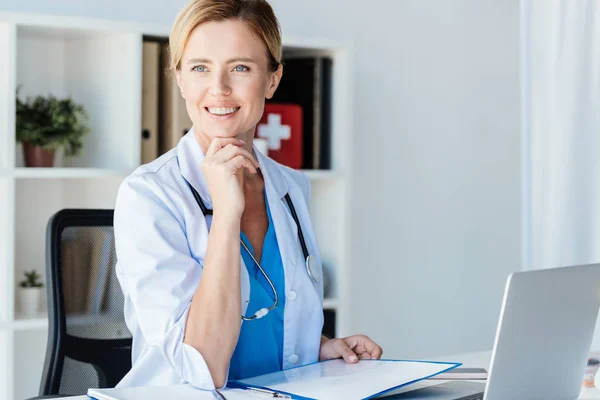 Doctora sonriente con estetoscopio mirando a la mesa con portapapeles y laptop en la oficina - foto de stock