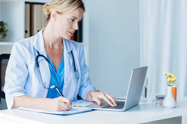 Doctora enfocada escribiendo en portapapeles en la mesa con computadora portátil en la oficina - foto de stock