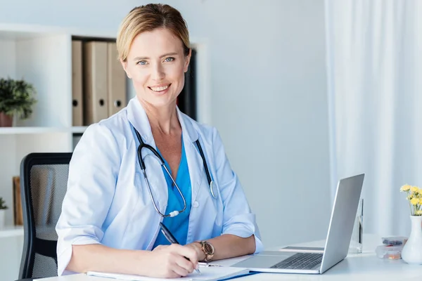Smiling female doctor looking at camera while writing in clipboard at table with laptop in office — Stock Photo