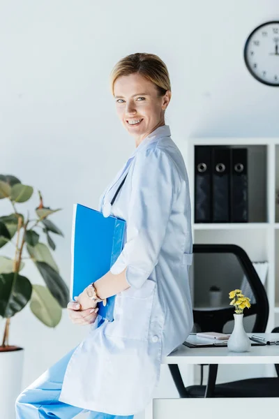 Sorridente medico femminile adulto con appunti seduto sul tavolo e guardando la fotocamera in ufficio — Foto stock