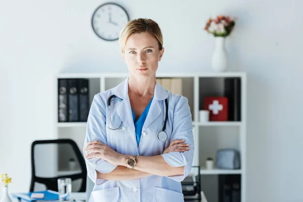 Femme médecin grave en manteau blanc avec stéthoscope au-dessus du cou debout avec les bras croisés regardant caméra dans le bureau — Photo de stock