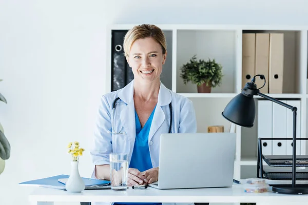 Smiling adult female doctor looking at camera and sitting at table with laptop in office — Stock Photo