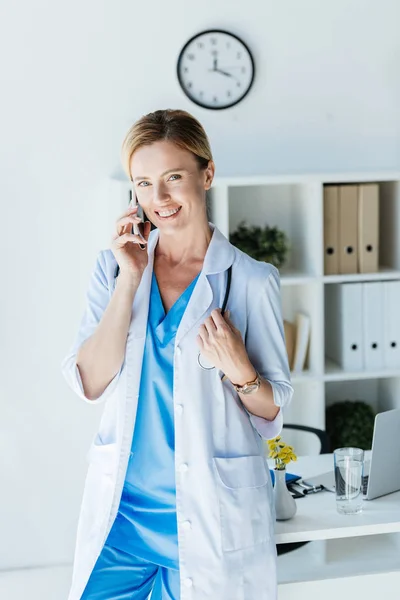 Приваблива жінка-лікар в білому пальто, дивлячись на камеру і розмовляючи на смартфоні в офісі — стокове фото