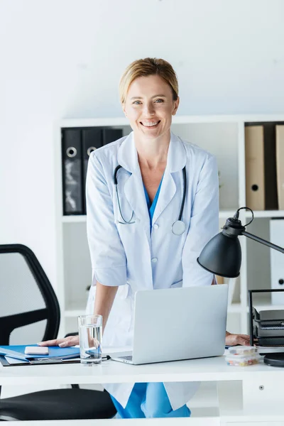 Mujer feliz médico en bata blanca con estetoscopio sobre el cuello de pie en la mesa con el ordenador portátil en la oficina - foto de stock