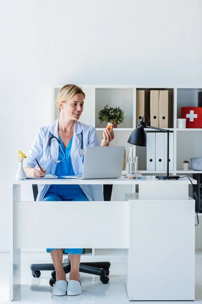 Médico feminino escrevendo e segurando pílulas à mesa com laptop no escritório — Fotografia de Stock