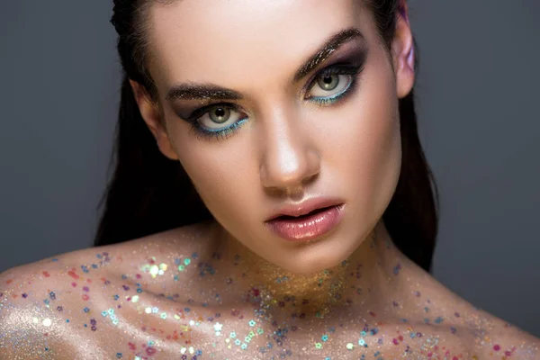 Glamorous girl with glitter on body and makeup looking at camera, isolated on grey — Stock Photo