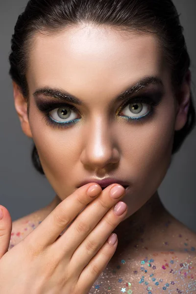 Chica glamorosa impactada posando con maquillaje y brillo en el cuerpo, aislado en gris - foto de stock