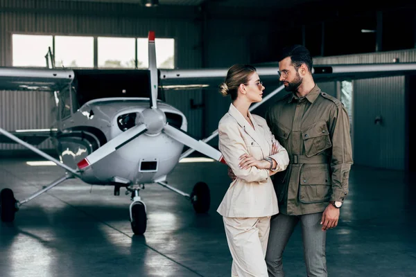 Élégant jeune couple en vestes se regardant près de l'avion dans le hangar — Photo de stock